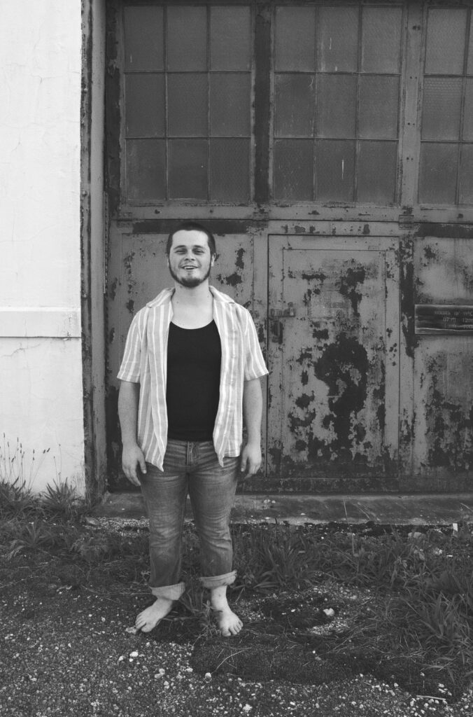 Tyler Nichols laughing barefoot in front of a weathered door, black and white
