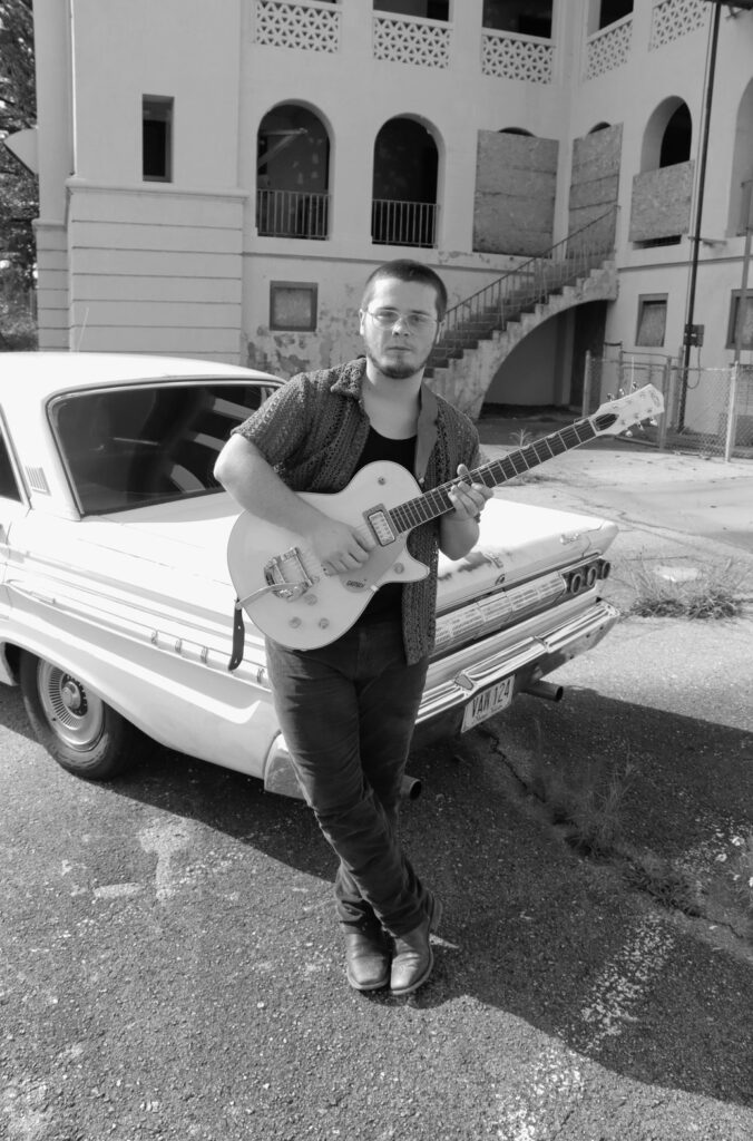 Tyler Nichols with a guitar beside a vintage car, black and white